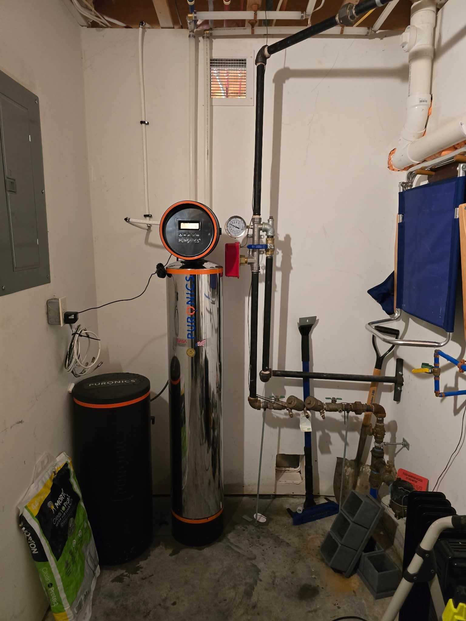 A Puronics tank with an orange control valve and brine tank installed on the main line of a home utility room, with copper and black supply piping, a pressure gauge, and shutoff valves visible.
