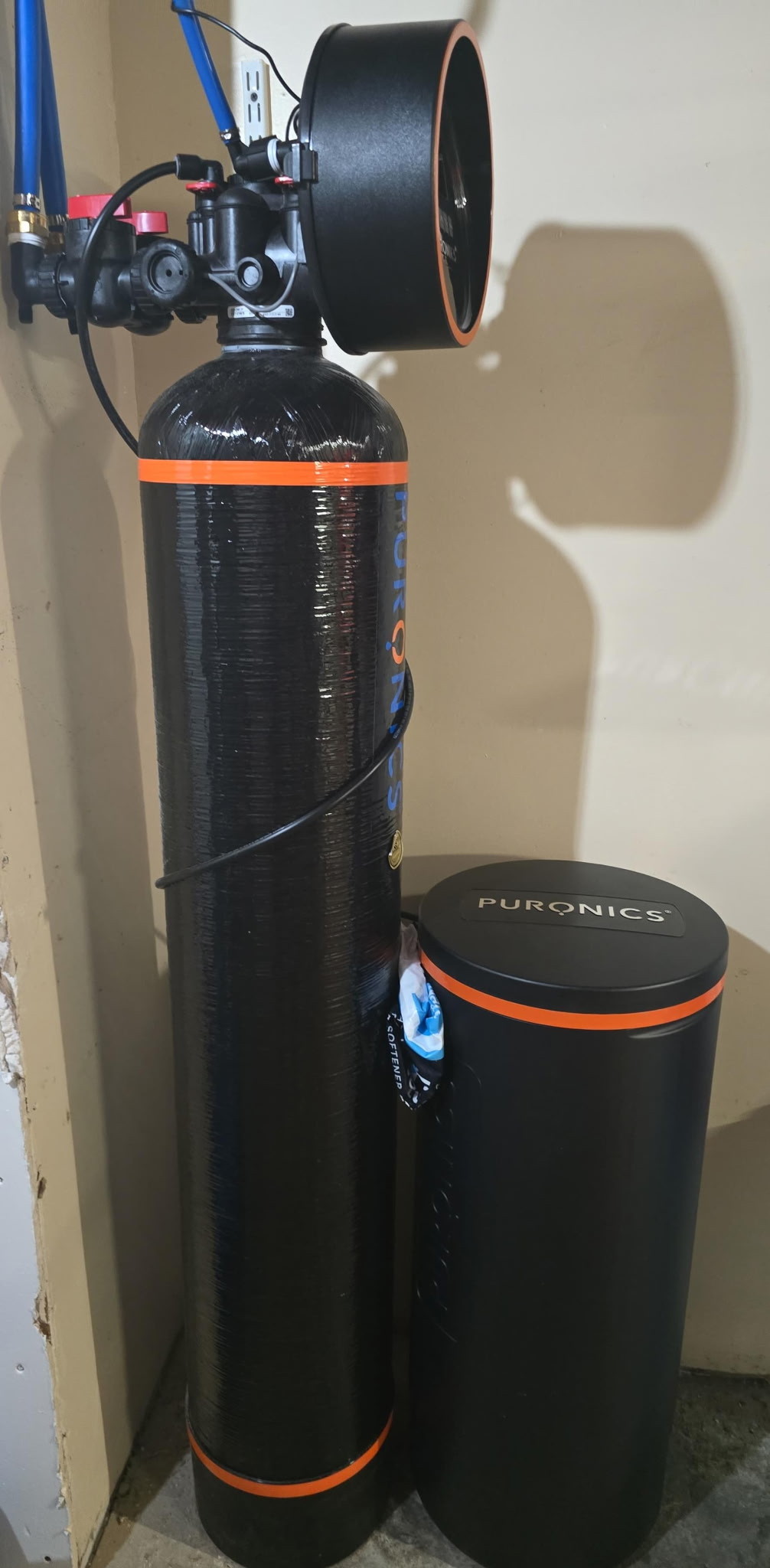 Close view of a black Puronics filtration tank with an orange band, control valve, and brine tank in a home utility area.